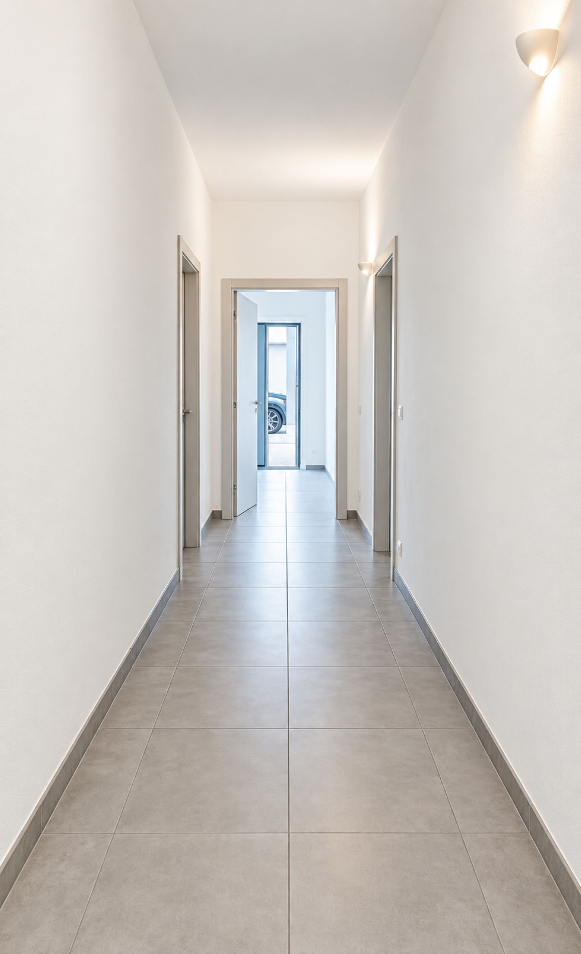 A corridor with a tiled floor and white walls, leading to a door at the end.