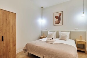 Une chambre moderne avec une armoire en bois, un lit recouvert d’une couvre-lit beige, deux tables de chevet avec des lampes et une œuvre d’art abstraite encadrée accrochée au mur.