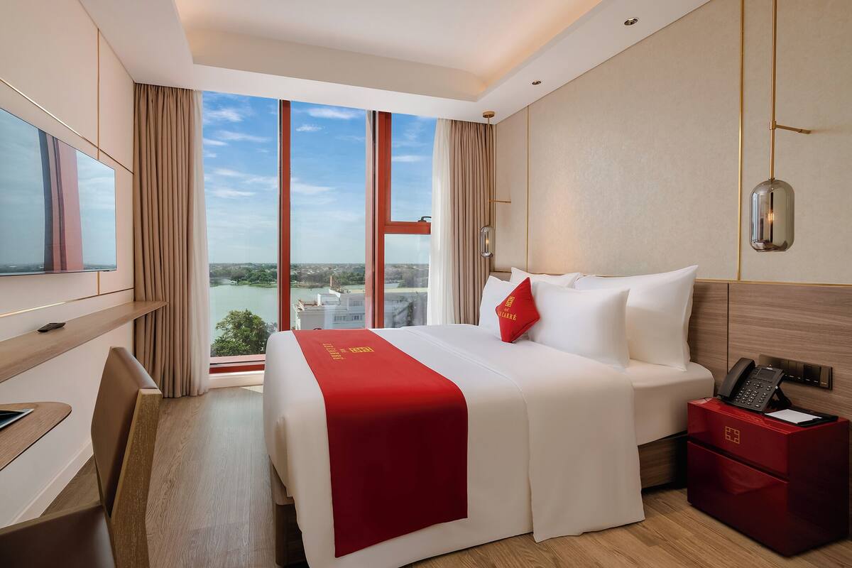 A hotel room with a large bed, a desk, a TV, and a view of a river and cityscape.