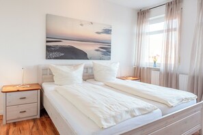 Un lit double avec une literie blanche, une table de chevet avec une lampe, une petite table de nuit avec un tiroir et une fenêtre avec des rideaux légers.