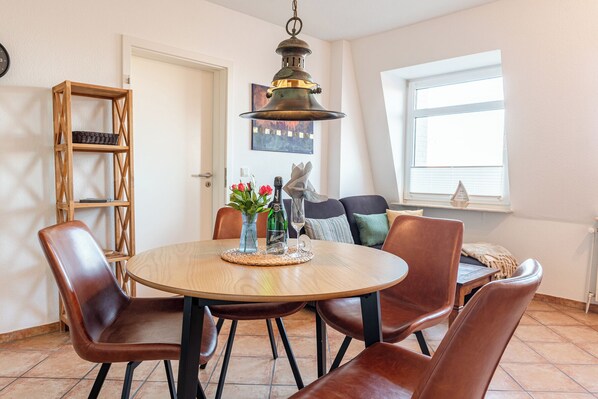 Un coin repas avec une table ronde, quatre chaises, un vase de fleurs, une bouteille de vin et une lampe décorative suspendue.