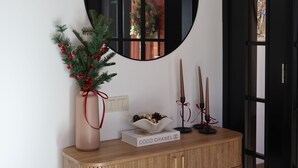 Una consola de madera con un espejo redondo encima, un florero con follaje, un candelabro y una cesta con un cojín.