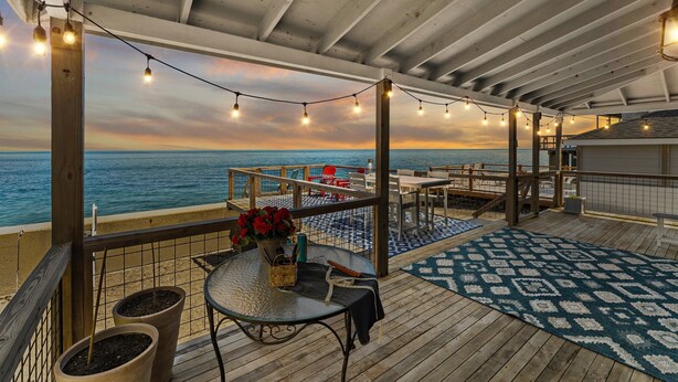 Um deck coberto ao ar livre com vista para o oceano, luzes de fadas, uma mesa com um vaso de flores e um tapete estampado.