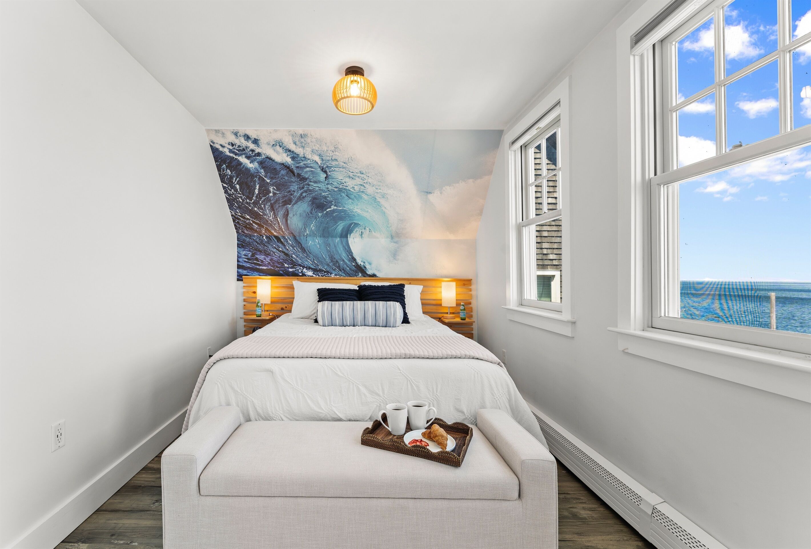 Quarto com um mural de uma grande onda do oceano, uma cama com roupa de cama branca, uma mesa de cabeceira com abajur e uma janela com vista para o mar.