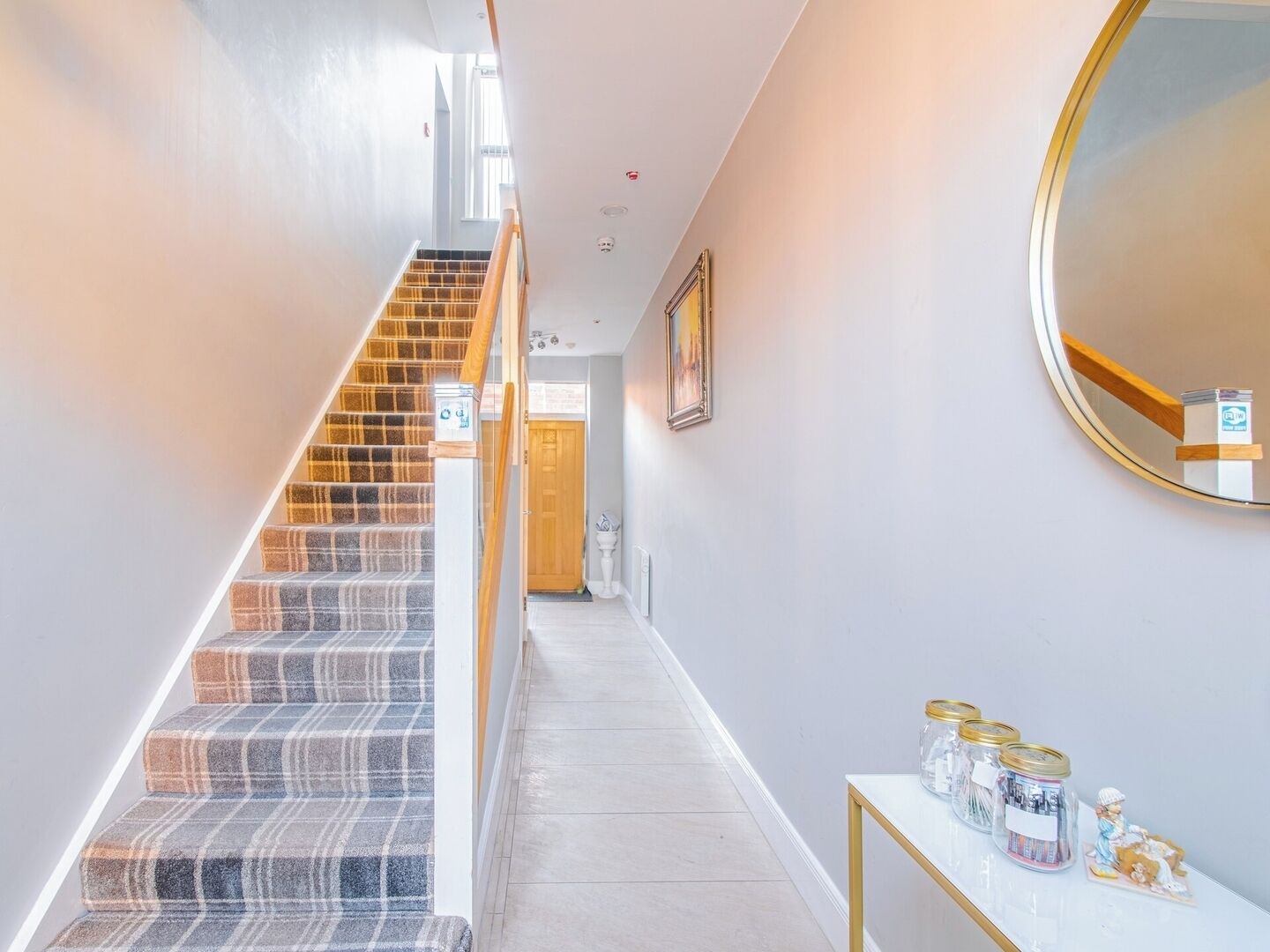 A staircase with a plaid carpet, a white wall, a round mirror, and a small table with jars.