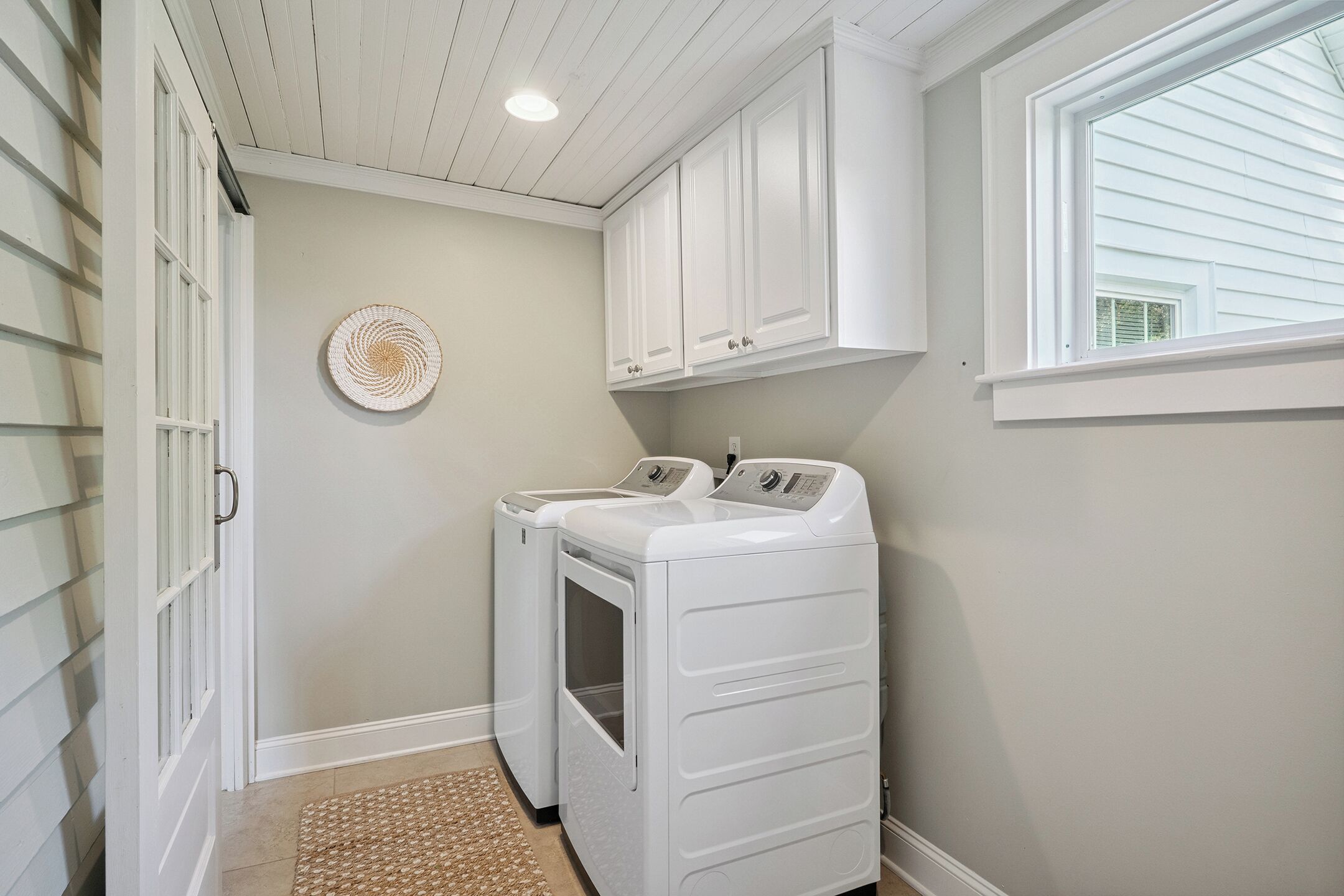 A laundry room with two white front-loading washers and dryers, a woven wall hanging, and a window with blinds.