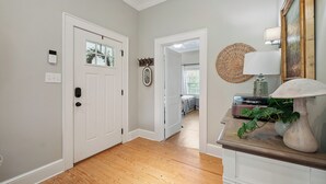 A hallway with a wooden floor, white walls, and a door with a window. There is a coat rack with items and a lamp on a table. - Bluebird Cottage in heart of Hickory, NC (Hickory)
