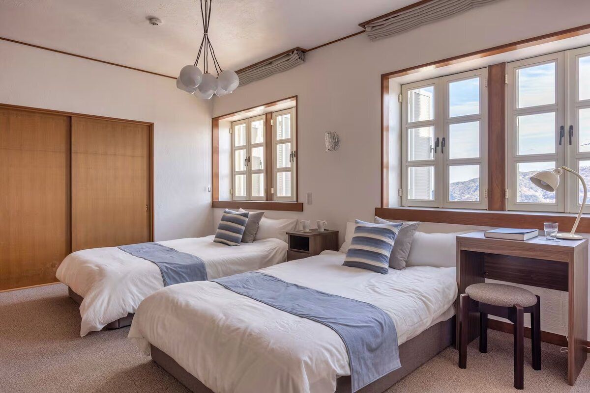 A bedroom with two beds, a desk, and a chair. There are windows with blinds, a ceiling fan, and a wall-mounted light fixture.
