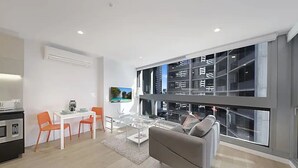Un salon moderne comprenant un canapé, une table à manger et des chaises, un coin cuisine équipé d’appareils électroménagers, et de grandes fenêtres offrant une vue sur la ville.