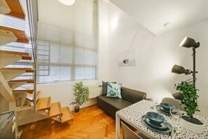 Een modern appartement met een houten trap, een zithoek met een bank, een eettafel gedekt voor twee personen en een plant in een pot.