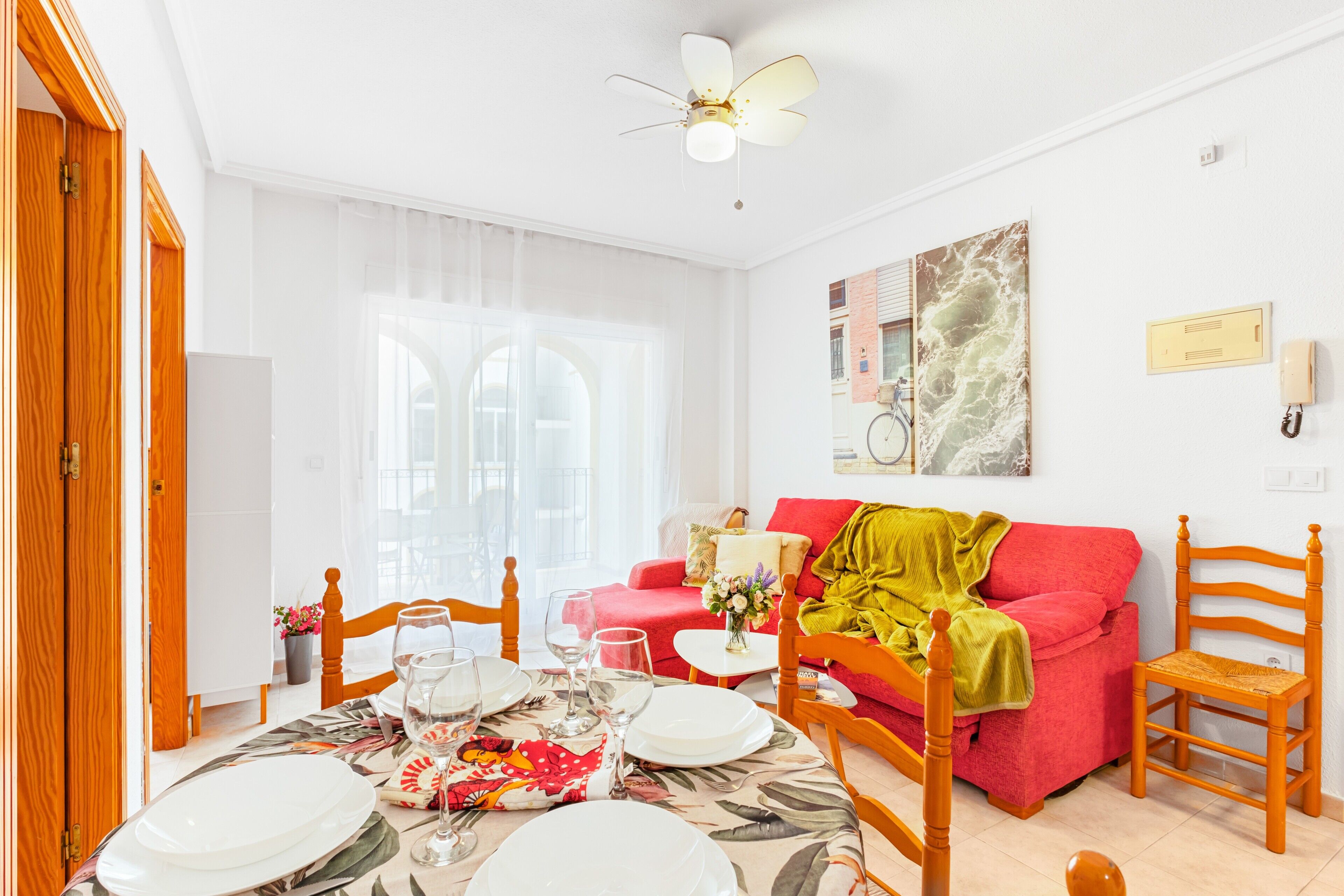 Un comedor con una mesa dispuesta para cuatro personas, un sofá rojo, un ventilador de techo y un teléfono fijo en la pared.