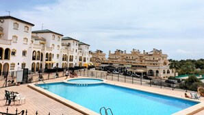 Une piscine entourée d’une terrasse carrelée, elle-même bordée de bâtiments blancs dotés de balcons et de fenêtres en arc de cercle.