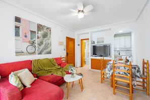 A living room with a red sofa, a small table with a vase, a dining area with a table and chairs, a TV, and a ceiling fan. - Apartment 'Buen Ambiente' with Shared Pool, Shared Garden and Wi-Fi (Orihuela)