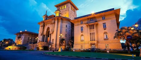 A large, historic building with a central tower and multiple wings, surrounded by a well-maintained lawn and palm trees.
