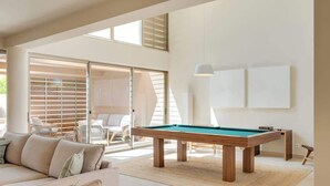 Ein modernes Wohnzimmer mit Billardtisch, einer Couch und einem großen Fenster mit Jalousien.