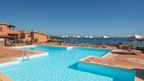 Ein Pool mit klarem, blauem Wasser, umgeben von Liegestühlen und Sonnenschirmen, mit Blick auf das Meer und ferne Berge.