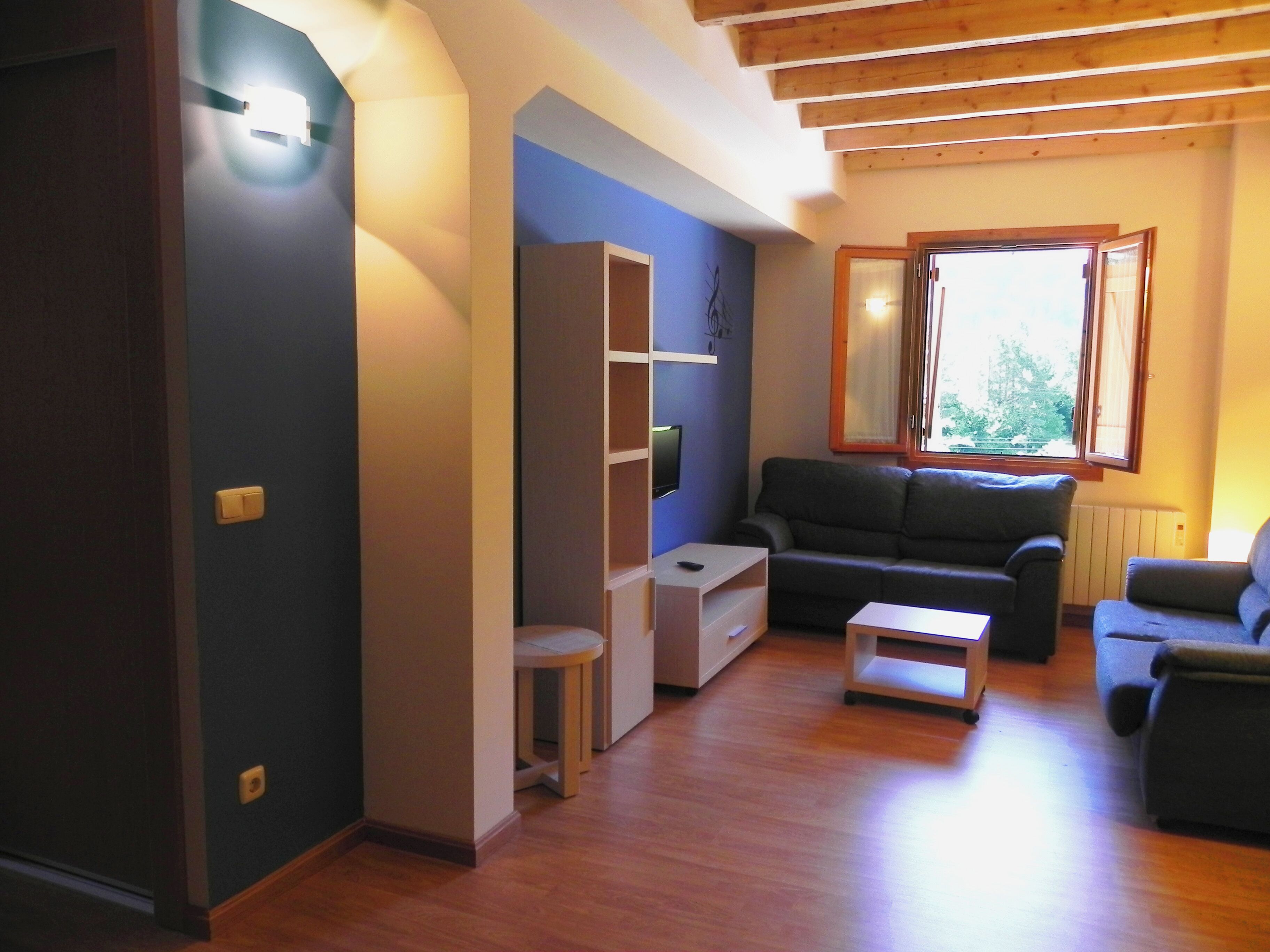 Sala de estar con pared azul oscuro, suelo de madera, sofá gris, una mesita blanca y ventana con vista a árboles.