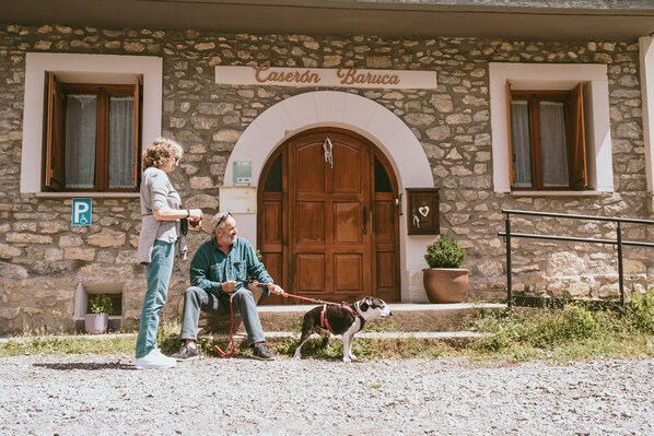 A stone building with a wooden door and a sign above it. There is a small potted plant on the ground. - Caserón Baruca (Bielsa)