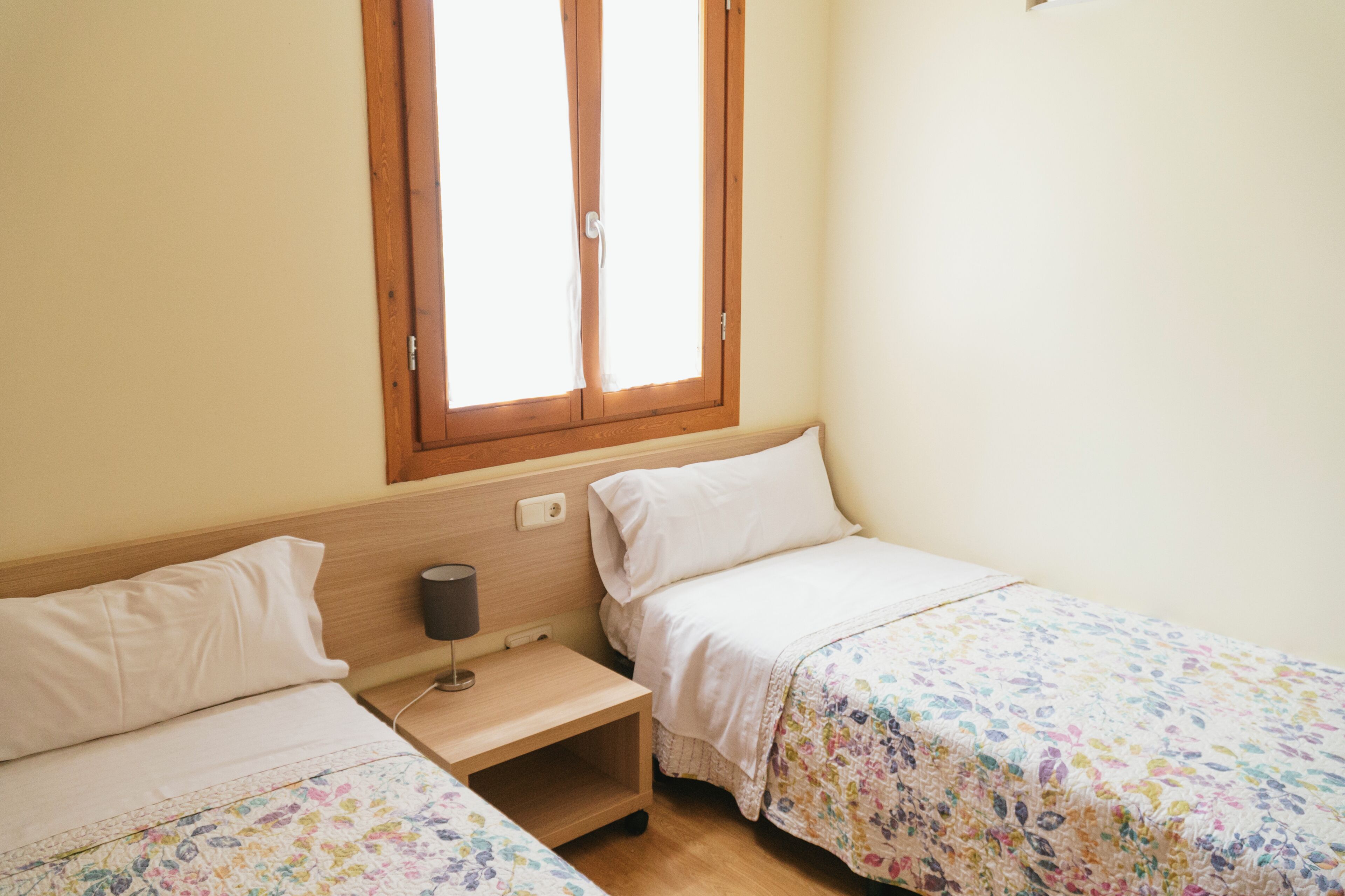 A small, clean room with two single beds, a nightstand with a lamp, and a window with curtains.