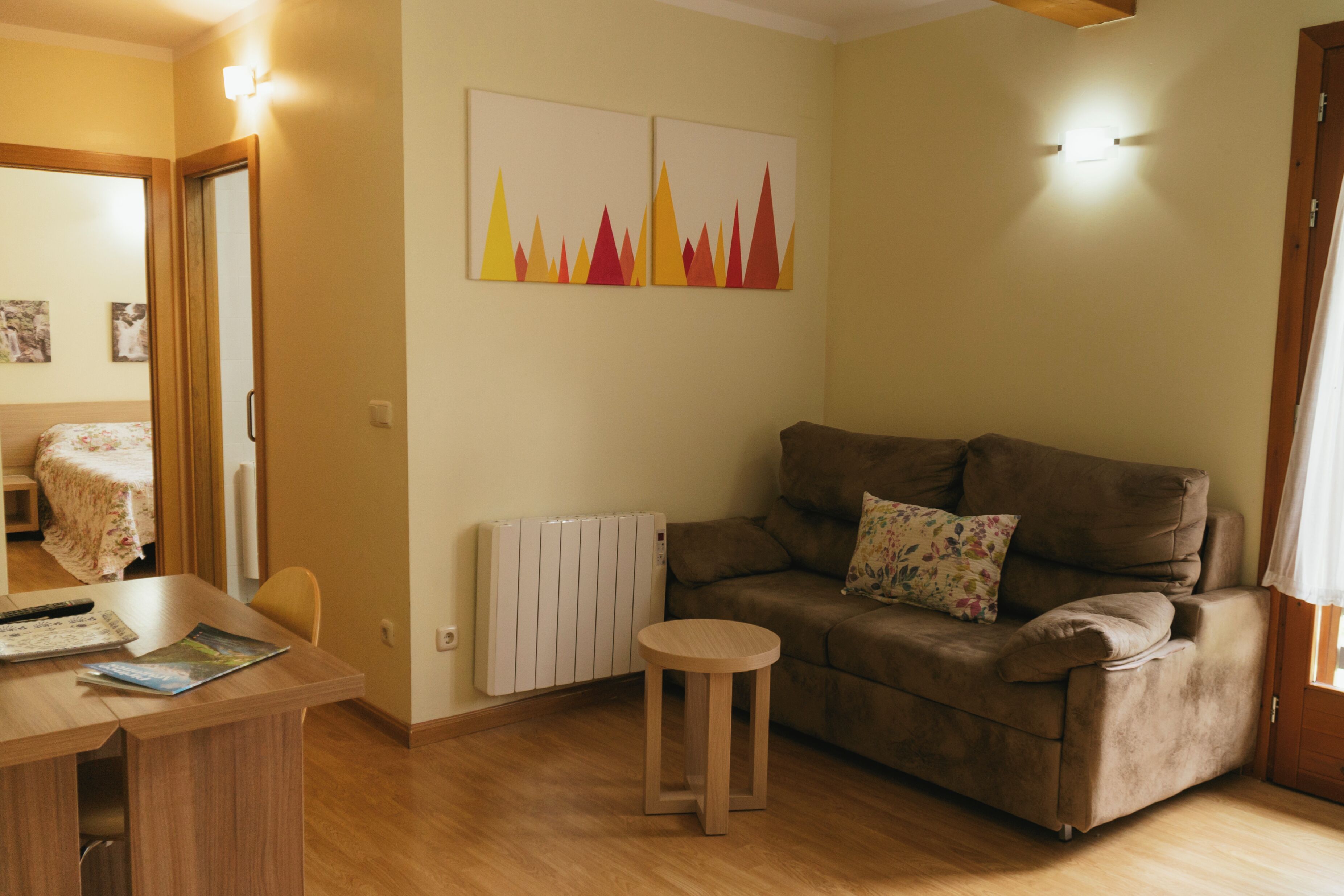 Una habitación con un sofá marrón, un taburete de madera, una mesa con un control remoto y una revista, y dos cuadros colgados en la pared.
