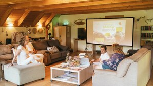 Ein gemütliches Wohnzimmer mit Holzboden, einer Projektorleinwand, die eine Präsentation zeigt, und einem Bücherregal voller Bücher.