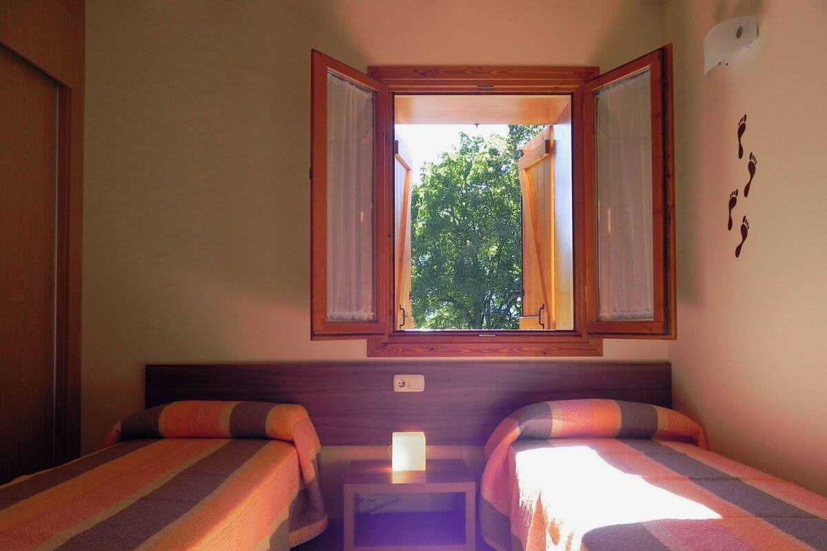 A room with two beds, a window with curtains, a small table, and a wall with a footprint decal.
