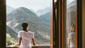 Eine Person steht an einem Fenster mit Blick auf eine gebirgige Landschaft.