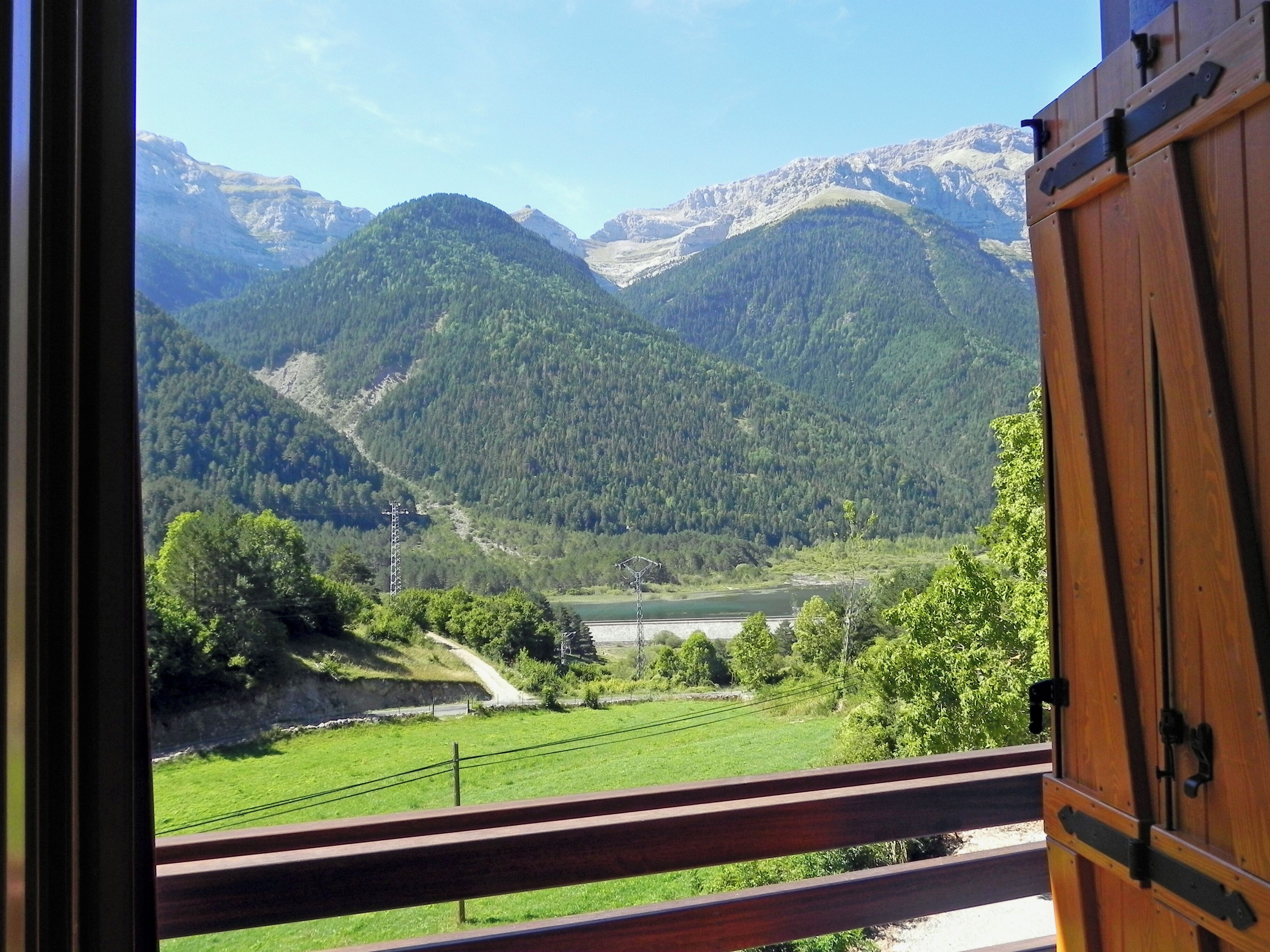 A view from a balcony with a wooden railing, overlooking a valley with mountains, a river, and a green field.