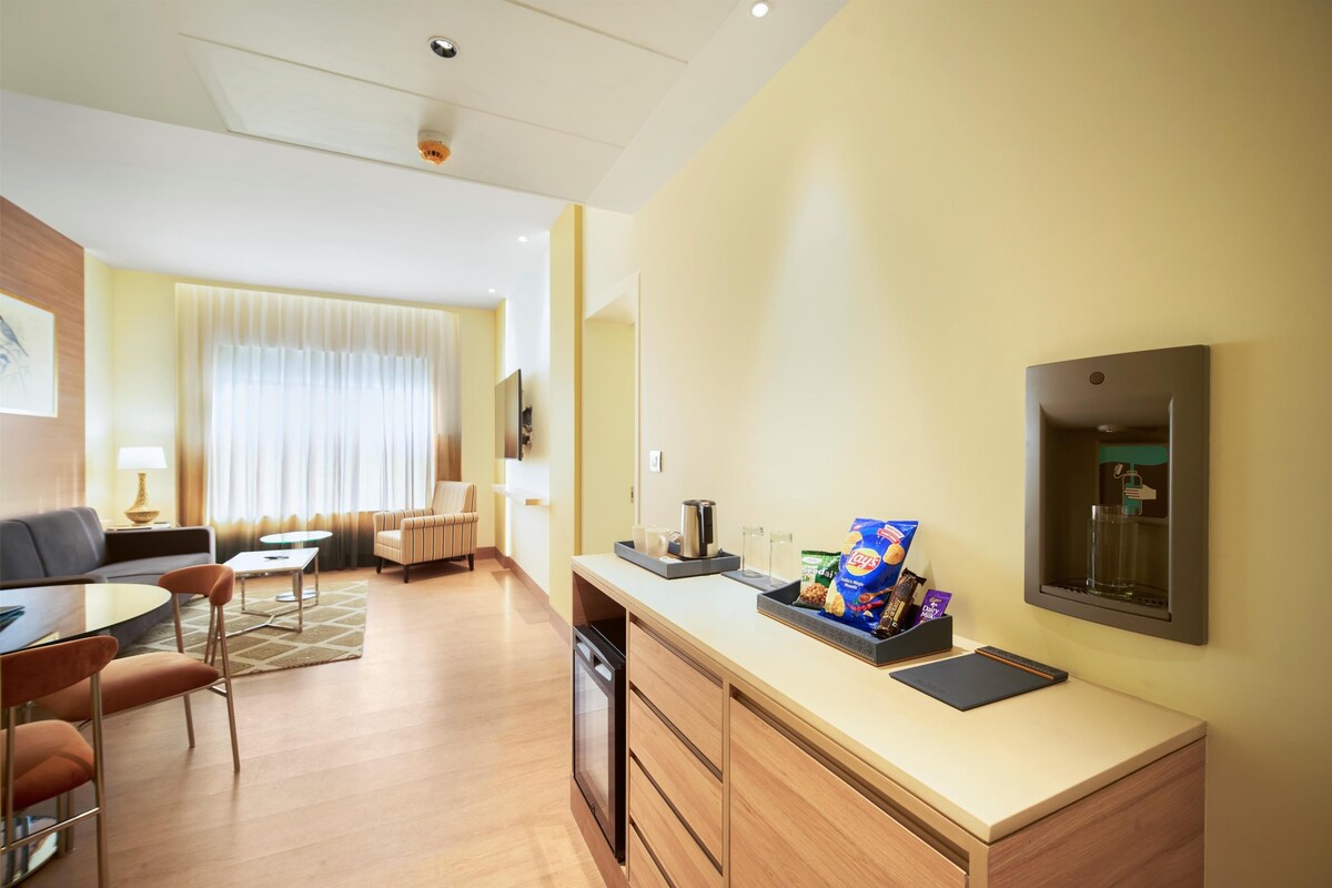 A modern hotel room with a dining area, a sofa, a coffee table, a TV, and a minibar.