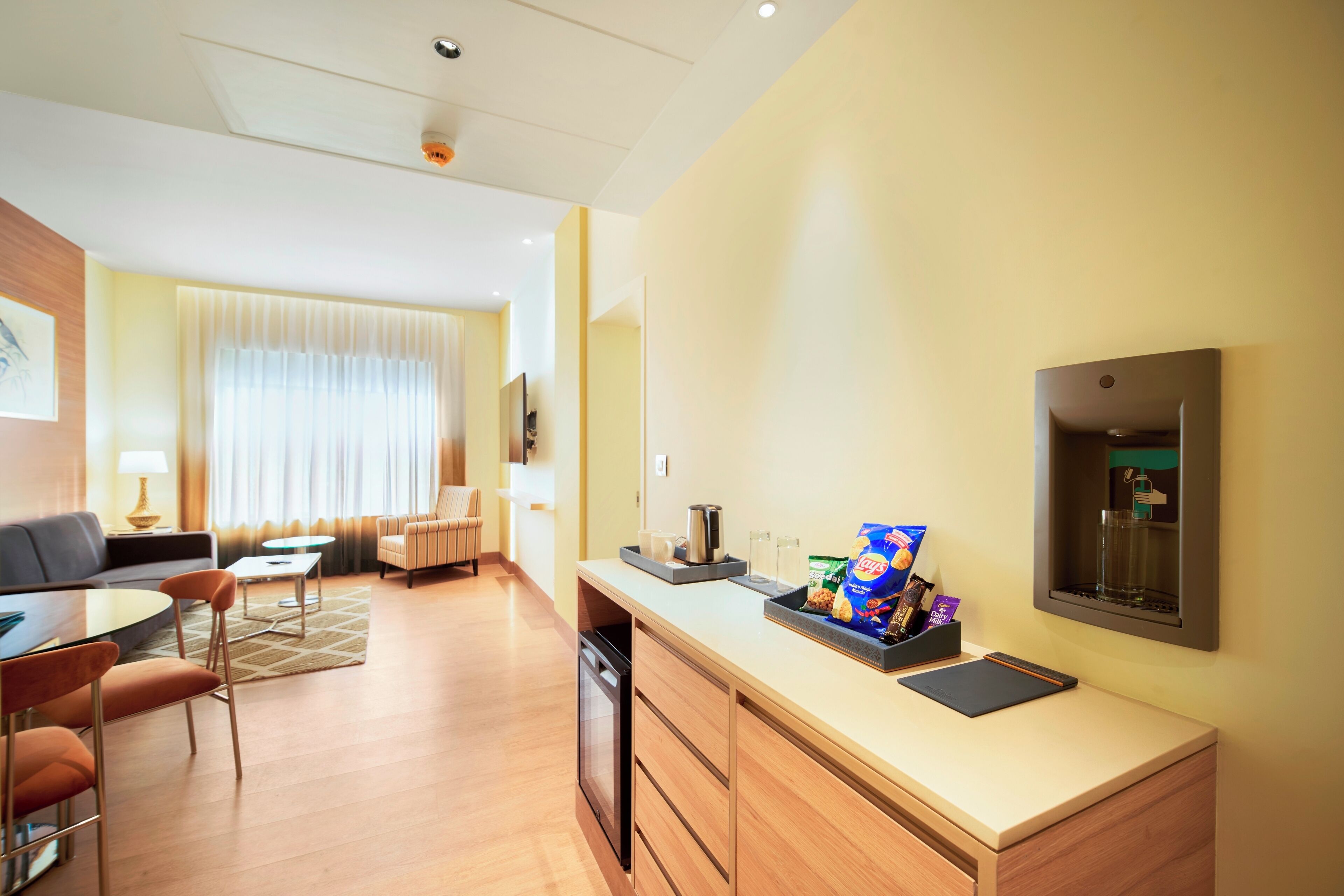 A hotel room with a dining area, a sofa, a coffee table, a chair, a desk with a computer, and a minibar.
