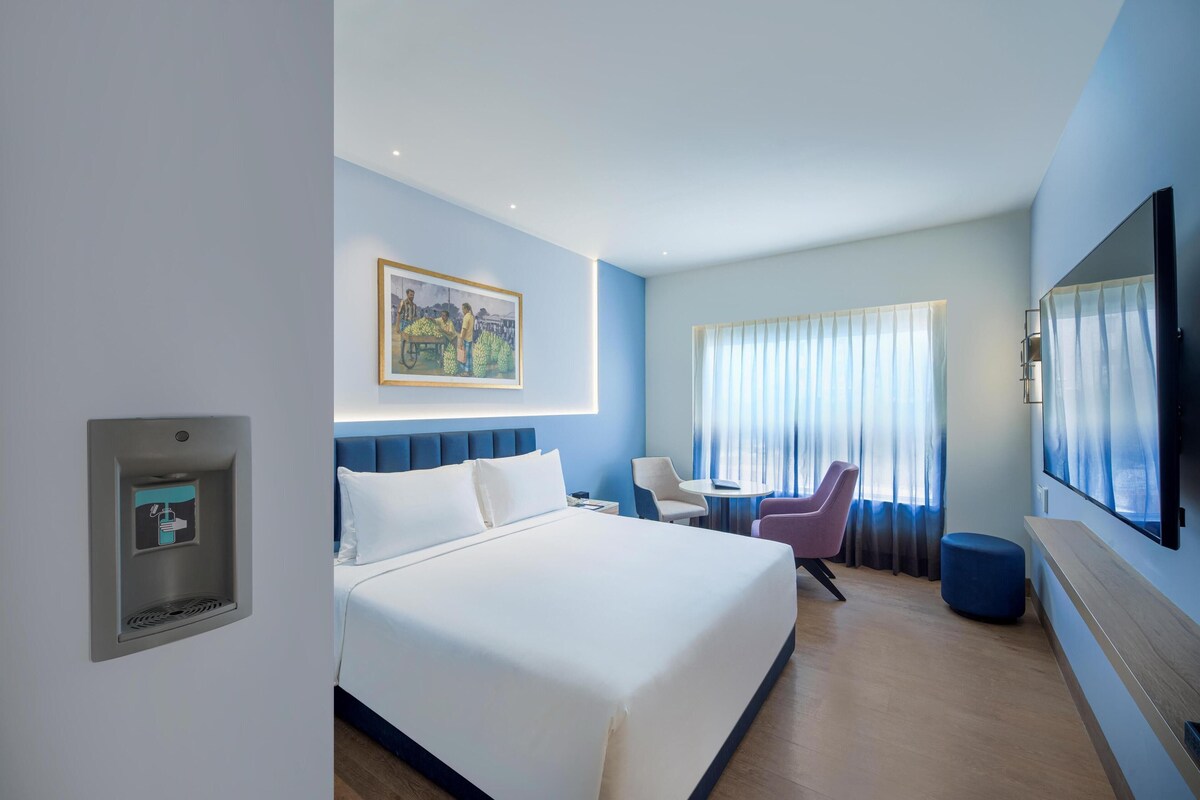 A hotel room with a bed, a desk, a chair, a TV, and a painting on the wall.