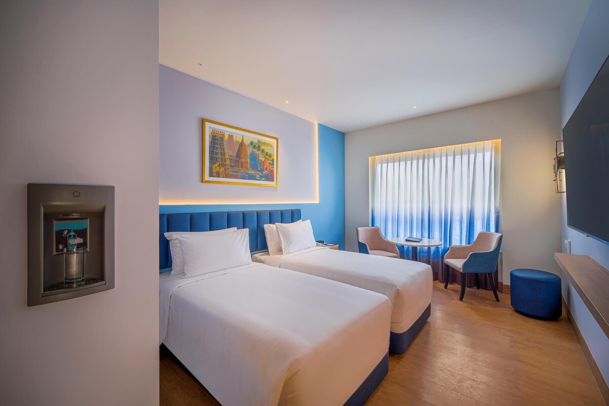 A hotel room with two beds, a water dispenser, a painting, a window with curtains, a small table, and a chair.
