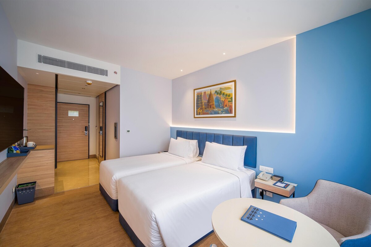 A hotel room with a large bed, a desk with a chair, a painting on the wall, and a blue accent wall.