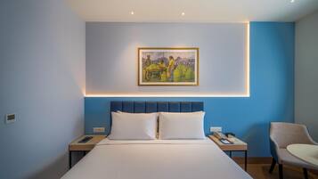 Una habitación de hotel con una cama grande, una mesita de noche con un teléfono y un libro, una silla y un cuadro en la pared.