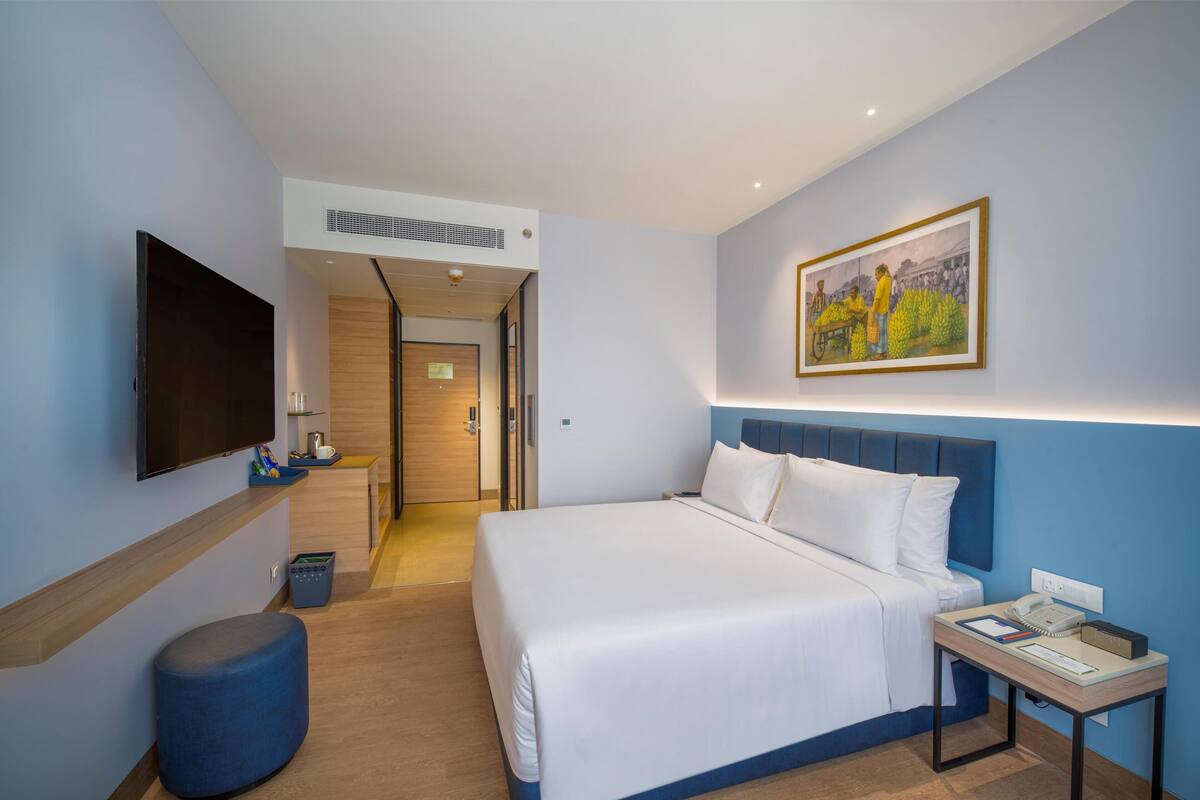 Une chambre d’hôtel avec un grand lit, un téléviseur à écran plat, une table de chevet avec un téléphone et un livre, un tableau accroché au mur et un petit tabouret.
