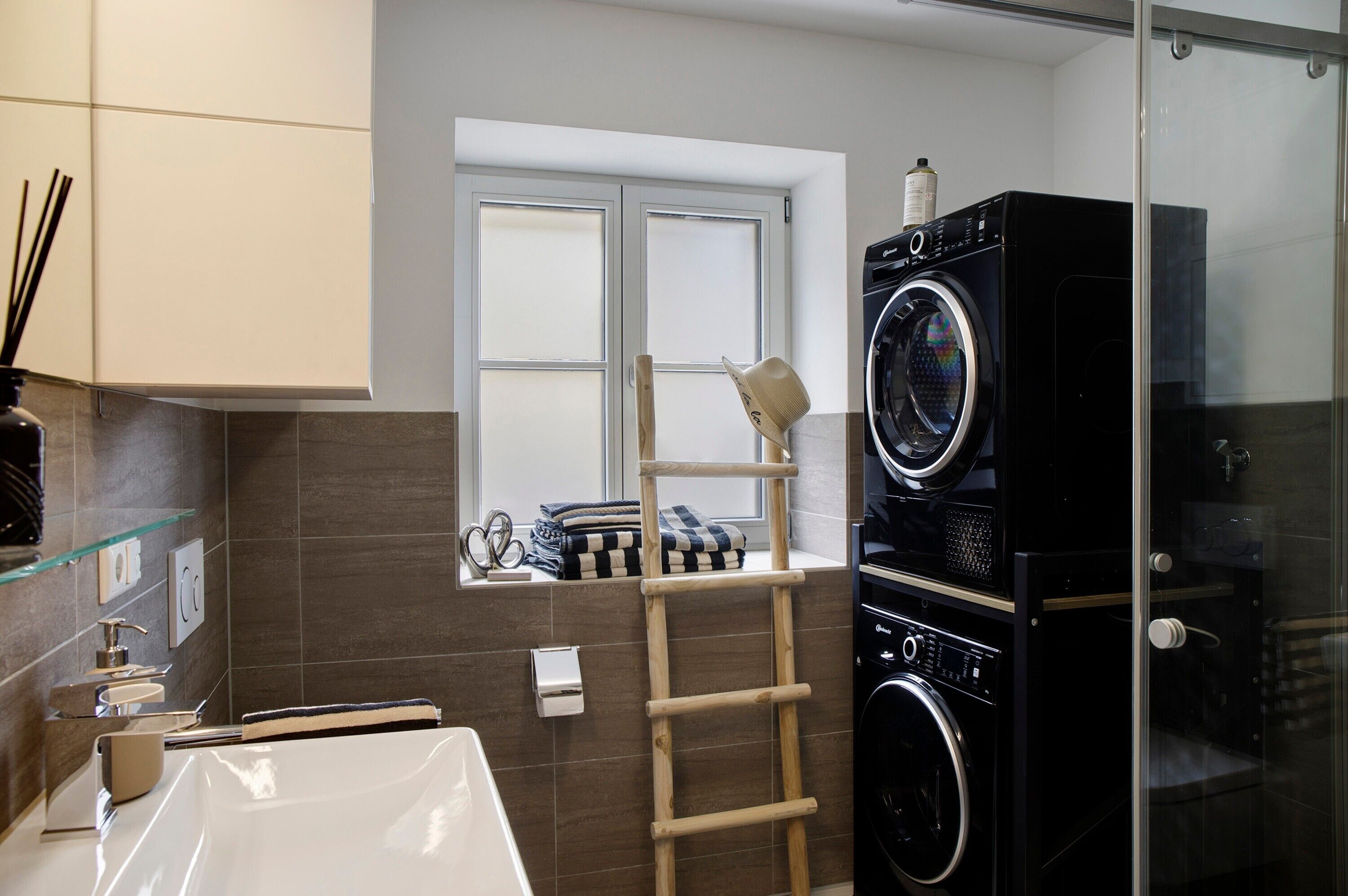A modern bathroom with a glass shower enclosure, a built-in washing machine, and a window with a view.