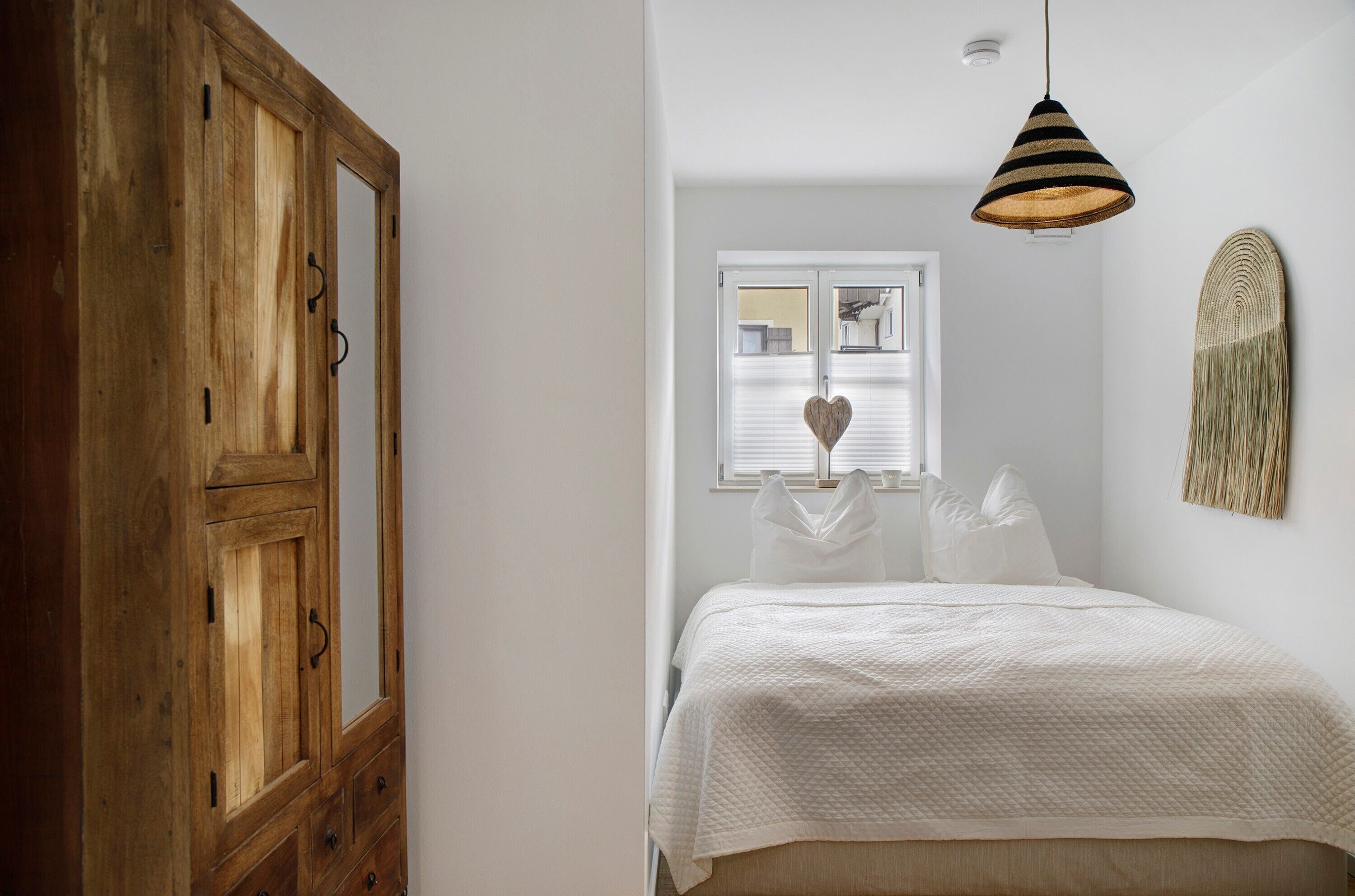 Ein Schlafzimmer mit einem Bett, einem Holzschrank, einer Pendelleuchte, einem Fenster mit Jalousien und einem Wandbild.
