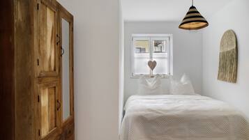 Ein Schlafzimmer mit einem Bett, einem Holzschrank, einer Pendelleuchte, einem Fenster mit Jalousien und einem Wandbild.