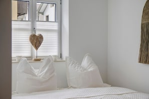 A bedroom with a bed, white pillows, a heart-shaped decoration on the window, and a woven wall hanging.
