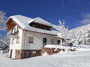 Un edificio de dos pisos con balcón, cimientos de piedra y alrededores cubiertos de nieve.