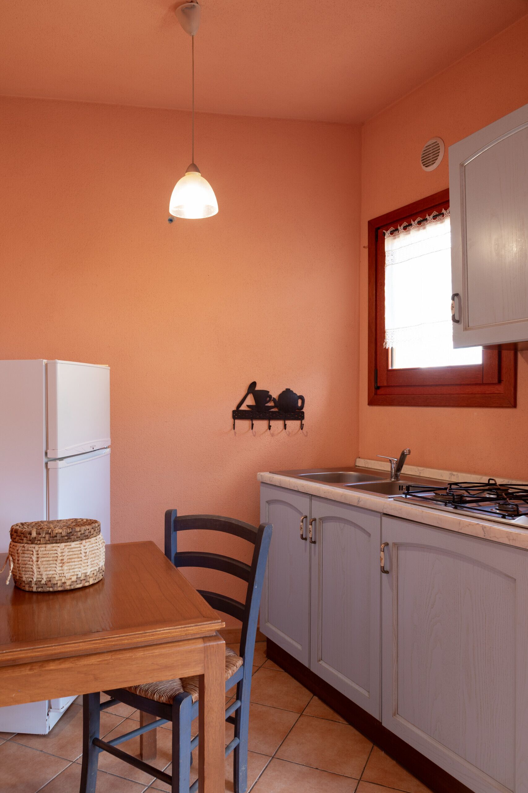 A compact kitchen with a refrigerator, a dining table with chairs, a hanging light fixture, and a window with a decorative valance.
