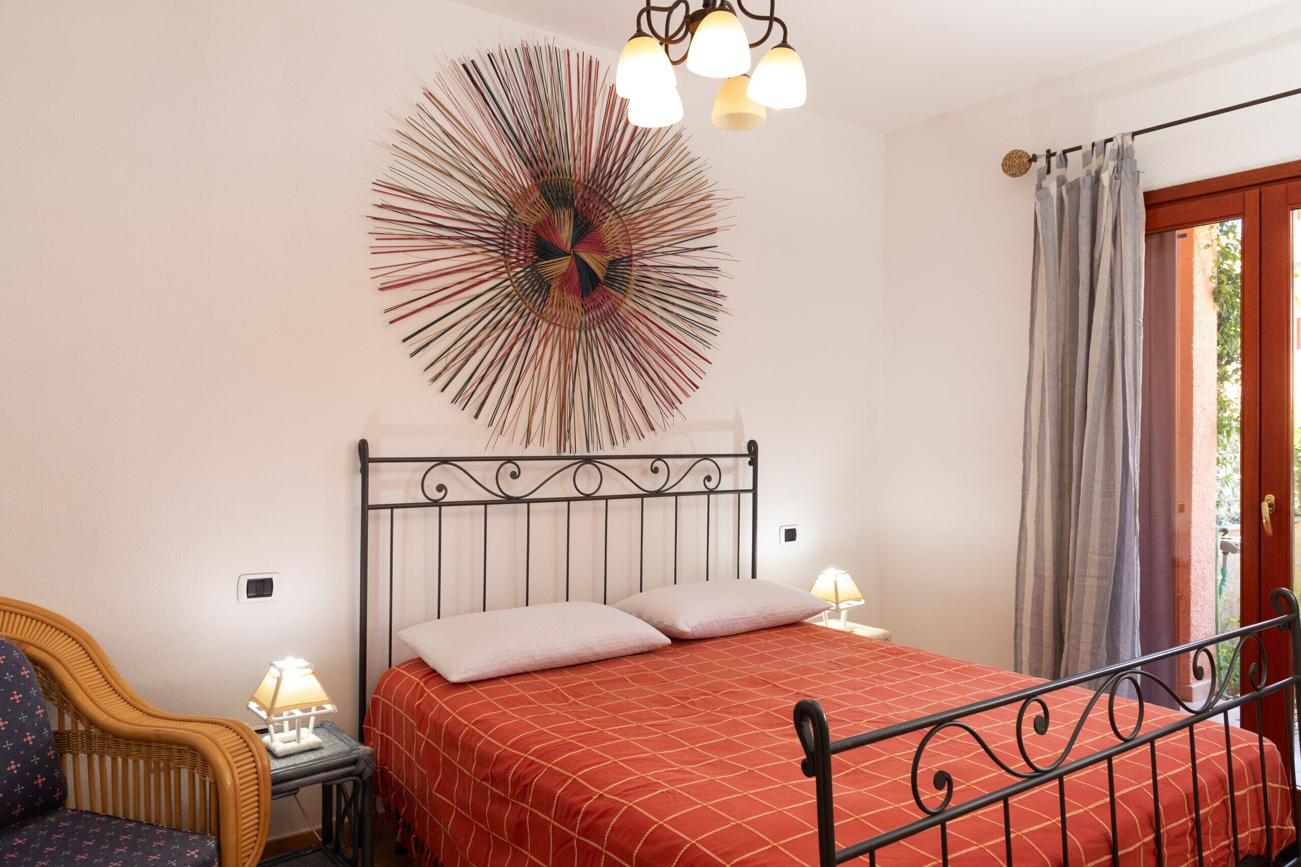 Um quarto com estrutura de cama de metal, colcha vermelha, uma obra de arte na parede, uma cadeira de vime e uma mesa de cabeceira com abajur.
