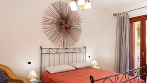 A bedroom with a metal bed frame, a red bedspread, a wall art piece, a wicker chair, and a bedside table with a lamp. - Casa Marzia by Wonderful Italy (Olbia)
