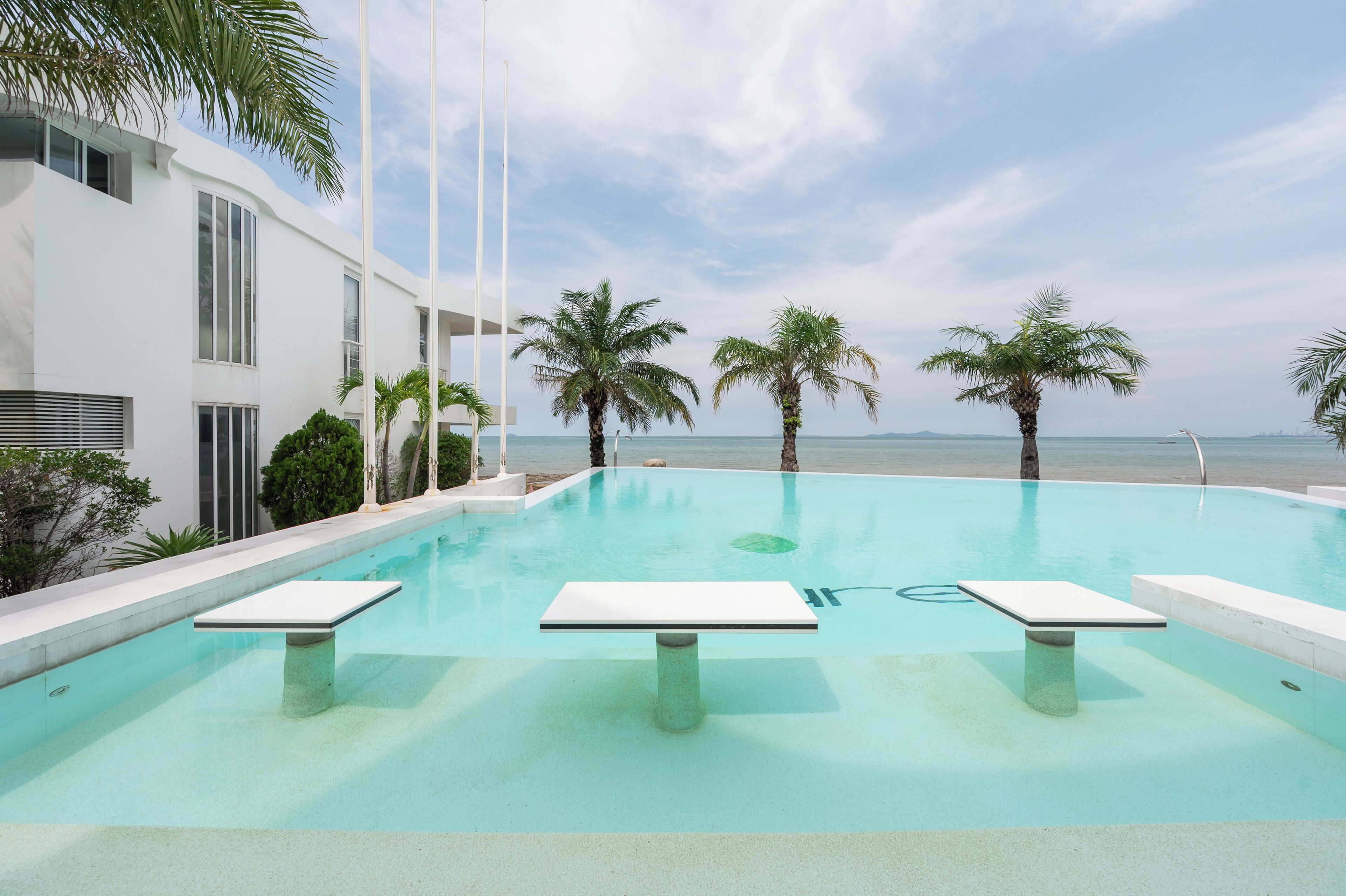A modern pool with a clear view of the ocean, surrounded by palm trees and a white building.