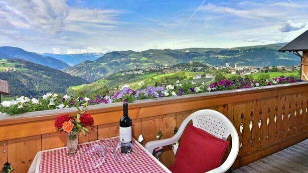 A table with a checkered tablecloth, a bottle of wine, and a vase of flowers on a balcony overlooking a valley.