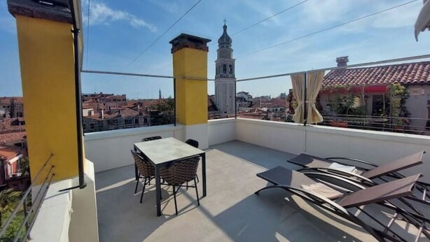 A rooftop terrace with a table and chairs, a yellow pillar, and a view of a cityscape including a church tower.