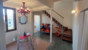 A modern living room with a glass table, a colorful sofa, a staircase, and a chandelier.