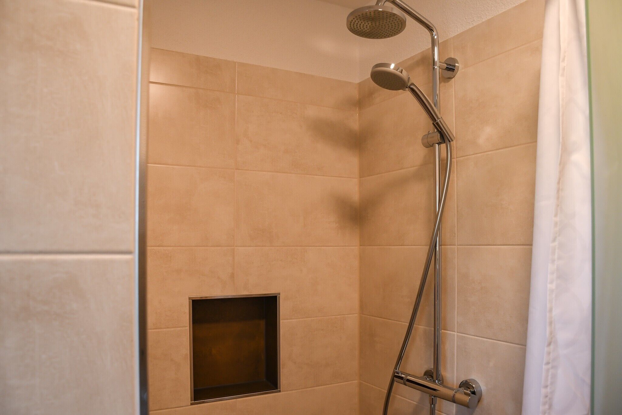 A shower with a handheld showerhead and a wall-mounted faucet, a small square vent, and a white shower curtain.