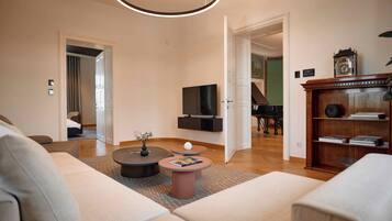 Una sala de estar moderna con un sofá, una mesa de centro y una televisión. Se observa un piano al fondo y una estantería con libros y un reloj.
