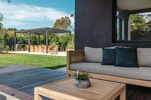 A modern outdoor seating area with a wooden table and a cushioned bench, a pergola, and a garden with potted plants.
